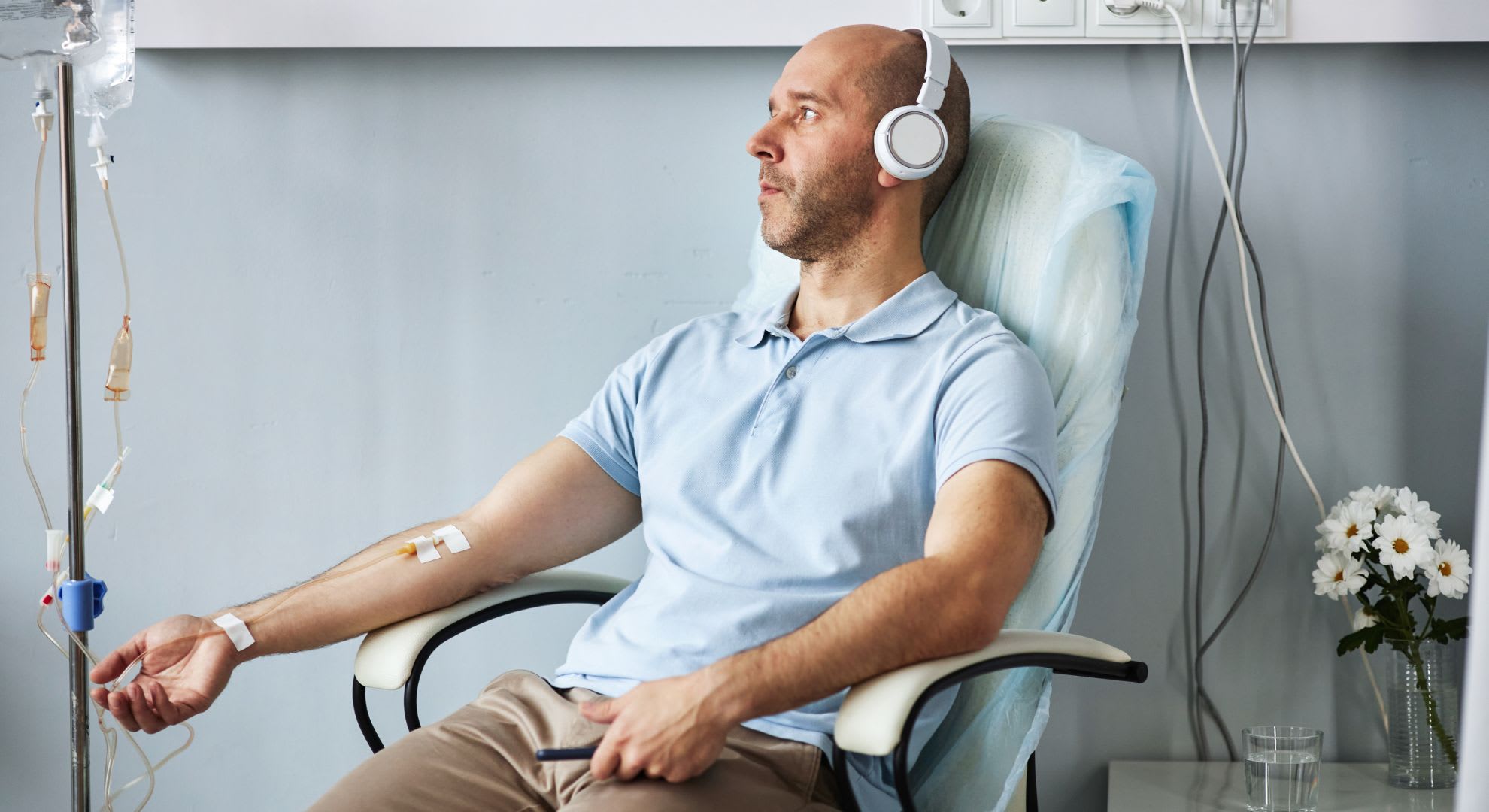 Bald man wearing headphones sits in a chair receiving intravenous therapy, highlighting the benefits of ozone therapy, with an IV drip attached to his arm and a table with flowers nearby.